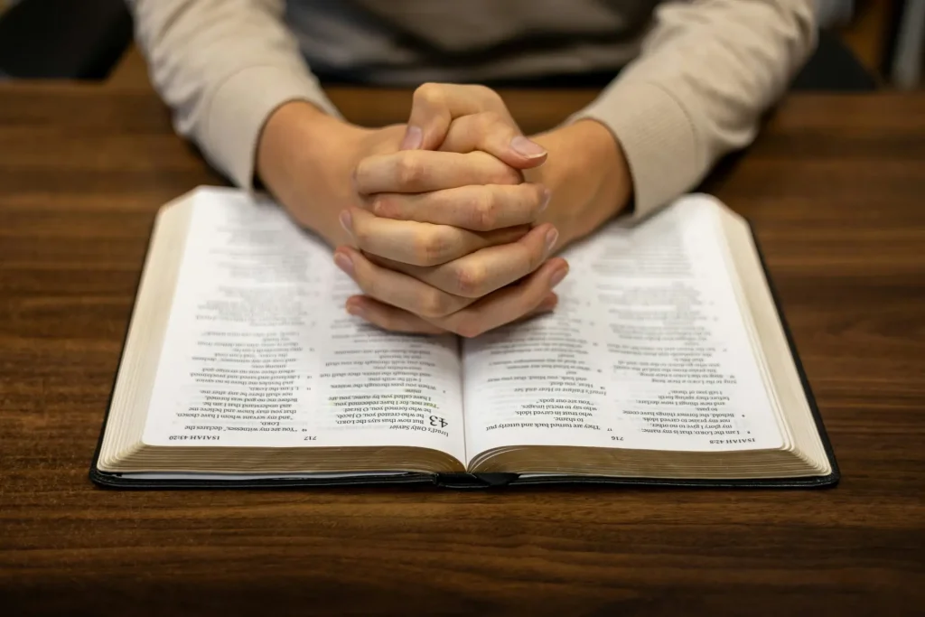 A person sitting at a table with hands folded over a book, symbolizing prayer and reflection on God's grace in difficult times.